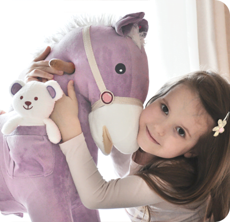 Uśmiechnięta dziewczynka przytula się do milutkiego konika bujanego. Konik ma z boku kieszonkę, w której siedzi biały pluszowy miś.