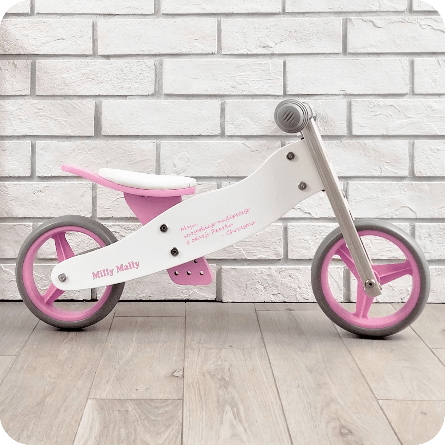 Rowerek biegowy dwufunkcyjny z personalizowany stoi na tle ściany z cegły. Posiada życzenia z okazji roczku.